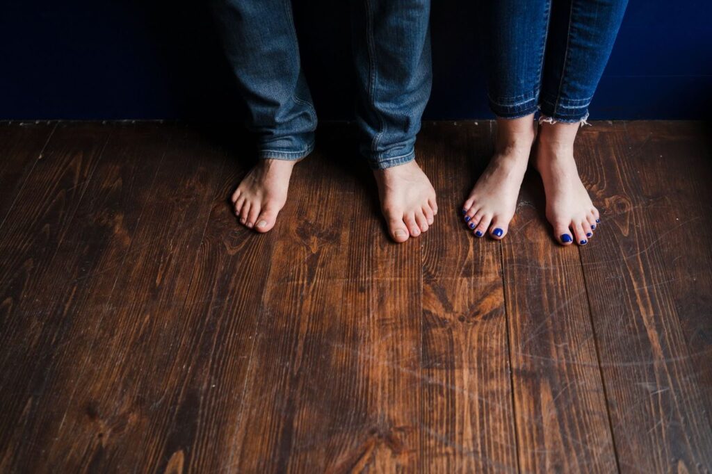Bare feet of a couple in blue jeans on warm-toned solid hardwood flooring.