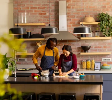Two people baking in a large kitchen with exposed brick features and a large island 